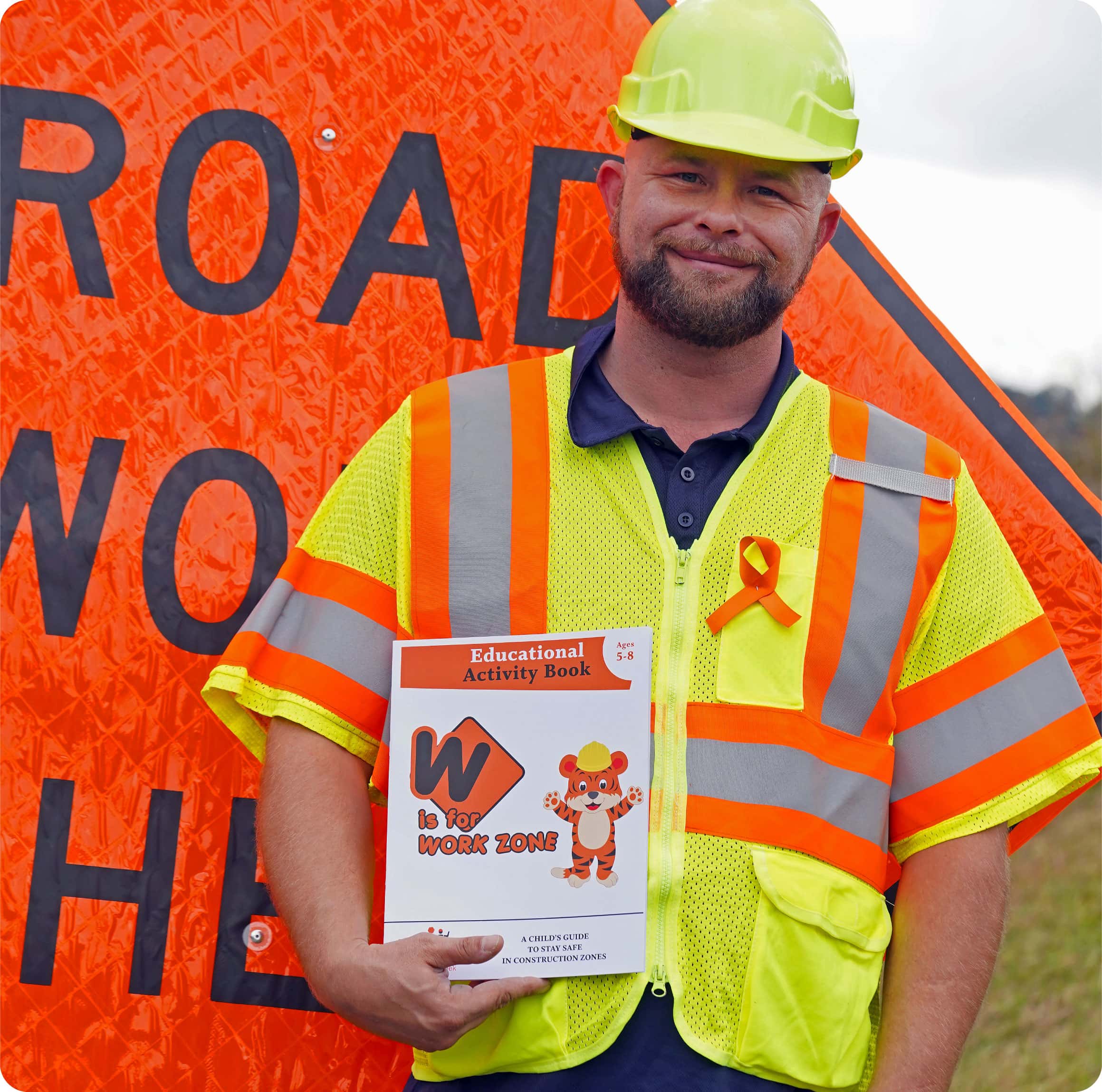 Roadway worker holding a copy of children's Work Zone Educational Activity Book while standing in front of a road sign